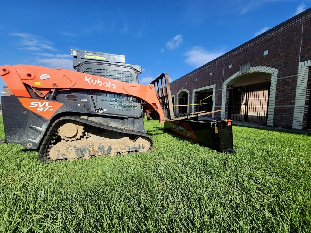 Kubota SVL97-2 compact track loader lifting a rack of pipes with a Ditch Witch attachment beside a brick utility building