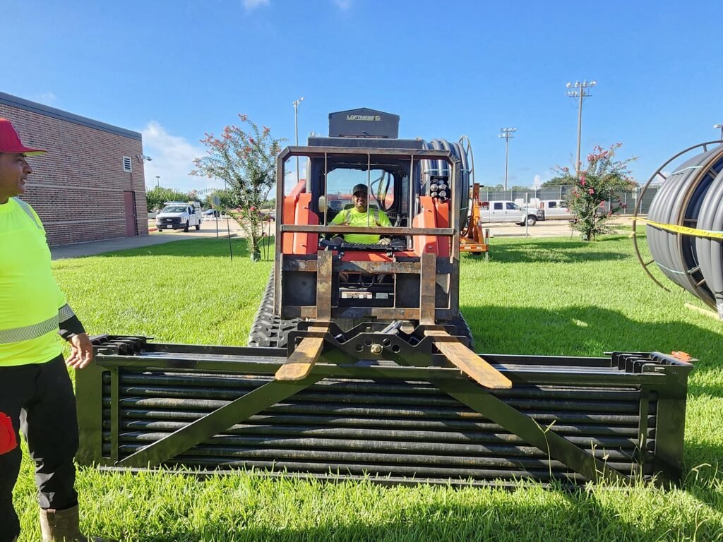 Utility workers staging a track loader with a fork attachment beside large conduit reels at an underground cable installation site.